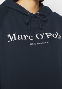 Granatowa bluza z kapturem wykonana z miękkiego materiału, z regulowanym sznurkiem przy kapturze. Na przodzie biały napis "Marc O’Polo EST. IN STOCKHOLM".
