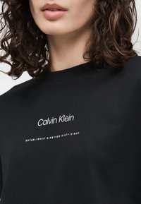 Γυναίκα με σγουρά σκούρα μαλλιά που φοράει μαύρο μπλουζάκι Calvin Klein με λευκό κείμενο «Calvin Klein» και «Established Nineteen Sixty Eight.»