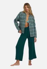 Groen geruite oversized shirt met twee borstzakken, gedragen over een patroon-bikini en teal wijde gebreide broek. Blote voeten, met minimale accessoires.