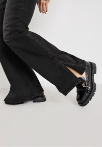 Mocasines gruesos negros con un acabado brillante en charol, que presentan un detalle de hebilla lateral y suelas de goma gruesas y texturizadas. Se llevan puestos unos vaqueros de denim negros acampanados.