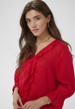 Pikkade pruunide juustega naine, kes kannab punast pluusi volangidega, kuldseid rõngaskõrvarõngaid, kihilisi kaelakeesid ja käevõru, poseerides valge tausta ees.