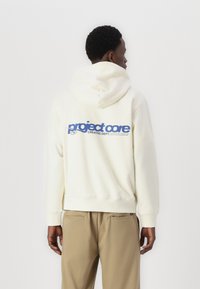 Sweat à capuche crème clair avec une poche kangourou, un grand graphique bleu avec l'inscription "project core" au dos, et des poignets côtelés aux manches.