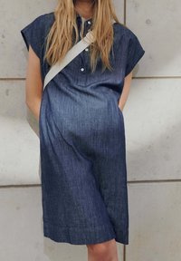Femme portant une robe en jean foncé à manches courtes boutonnée avec une bandoulière claire devant un mur carrelé clair.