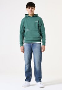 Felpa verde con tasca frontale e dettagli testuali, abbinata a jeans di denim blu e sneaker bianche. Texture liscia, design casual.