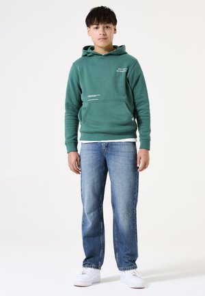 Felpa verde con tasca frontale e dettagli testuali, abbinata a jeans di denim blu e sneaker bianche. Texture liscia, design casual.