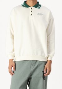 Osoba ubrana w kremową koszulę polo z długimi rękawami, zielonym kołnierzykiem i guzikami, w połączeniu z jasnzielonymi spodniami z czarną grafiką na udzie.