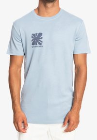 Muž v světle modrém tričku s krátkým rukávem a tmavě modrým abstraktním logem a textem "QUIKSILVER" na hrudi, kombinovaném s bílými kalhotami.