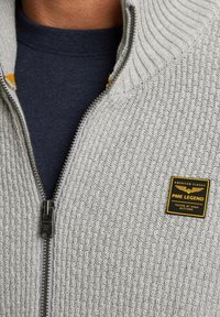 Grijze gebreide zip-up trui met een textuurpatroon, metalen rits, en een zwart-geel label op de borst. Daaronder zit een marineblauwe shirt.