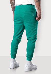 Grüne Sweatpants aus weichem Stoff mit schmal zulaufenden Beinen, ausgestattet mit einer Gesäßtasche und gerippten Bündchen. Kombiniert mit weißen Sneakers.