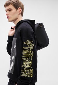 Μαύρο φούτερ με ριμπ υφή, με γραφικό στο μπροστινό μέρος και μεγάλα κίτρινα γράμματα στο μανίκι. Το μοντέλο φοράει μαύρο σακίδιο πλάτης.