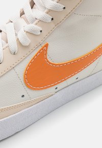 Nike-sneaker med en beige läderovandel, en ljusorange swoosh-logotyp med vit sömnad, och en vit gummisula.
