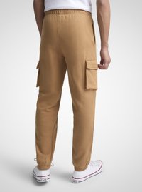 Brune bomullscargobukser med elastisk midjebånd, sidelommer og tettsittende mansjetter. Brukt med hvite Converse-sneakers.