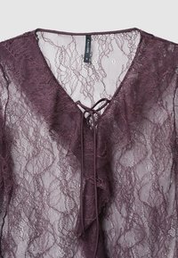 Top in pizzo di un profondo bordeaux con scollatura a V e bordi arricciati. Presenta un dettaglio annodato al collo e un intricato motivo floreale in pizzo.