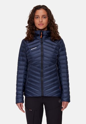 Vrouw in een marineblauwe gewatteerde jas met een hoge kraag en een ritssluiting aan de voorkant, gecombineerd met zwarte broek, staand tegen een witte achtergrond.