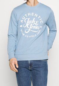 Světle modrá mikina s dlouhými rukávy, bílý potištěný nápis "AUTHENTIC Jack & Jones" a "DENIM CO SINCE 1990" ve tvaru kruhu.