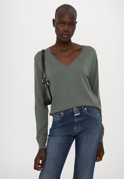 Suéter verde de punto con escote en V, de manga larga, combinado con jeans azul oscuro. La modelo lleva un pequeño bolso cruzado negro.