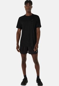 Schwarzes sportliches T-Shirt mit strukturiertem Design, kurzen Ärmeln und Logo. Kombiniert mit schwarzen Shorts. Modell trägt schwarze Laufschuhe.