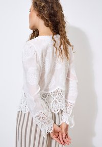 Wei�ße Spitzenbluse mit floralen Mustern, gewelltem Saum und langen Ärmeln. Ausführliche Häkelakzente an den Manschetten und am Saum. Gestreifte Hose sichtbar.