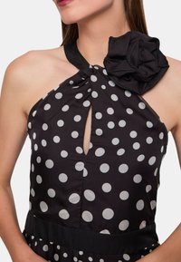 Abito halter nero con pois bianchi, caratterizzato da un'apertura a chiave e un grande fiore di tessuto decorativo sulla spalla. Texture liscia.