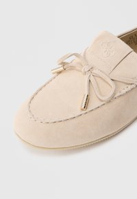 Μπεζ σουέντ μοκασίνια με στρογγυλεμένη μύτη, διακοσμητικό φιόγκο και ακροδέκτες σε χρυσό τόνο. Ραμμένη λεπτομέρ�εια κατά μήκος των άκρων. Ομαλή υφή.