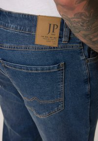 Denim jeans in dark blue with a brown leather patch labeled "JP 1880" on the waistband, featuring standard back pockets and visible stitching.