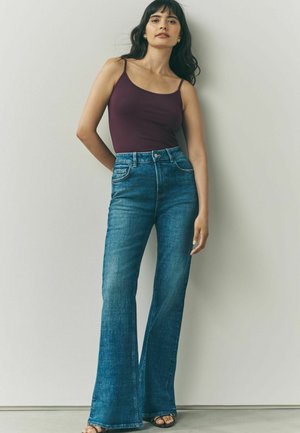 Hoogoplopende flared jeans in middelblauw denim, gecombineerd met een aansluitend bordeauxrood tanktop. Bevat vijf zakken en een subtiele textuur.