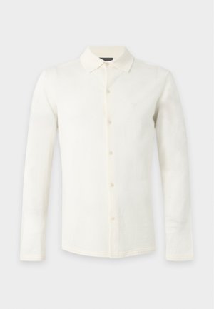 Jeune homme debout portant une chemise crème à manches longues, un jean bleu clair à jambes larges et des baskets blanches sur un fond blanc uni.