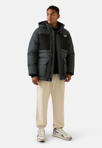 Donzen jas in donkergrijs met zwarte panelen, voorzien van ritszakken, een capuchon en een doorgestikt ontwerp. Draagt deze over een zwart shirt en beige broek.