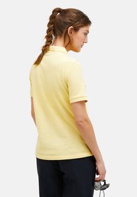 Polo shirt giallo chiaro, maniche corte, tessuto texturizzato, con colletto classico, abbinato a pantaloni scuri. Gli accessori includono occhiali da sole.