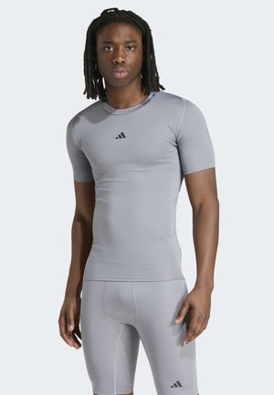 Mies, jolla on dreads, pukeutuu vaaleanharmaaseen istuvaan urheilupaitaan ja -shortseihin, joissa on mustat adidas-logot yksivärisen vaalean taustan edessä.