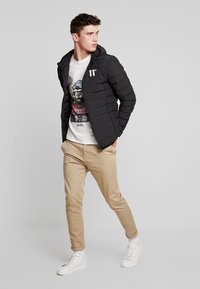 Chaqueta de plumas negra con capucha, con diseño acolchado; usada sobre una camiseta gráfica blanca. Pantalones caquis y zapatillas blancas completan el look.