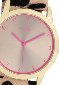 Gold-tone watch with a round face, pink hour markers, and hands. Beige strap with a black leopard print pattern. Smooth texture and modern design.