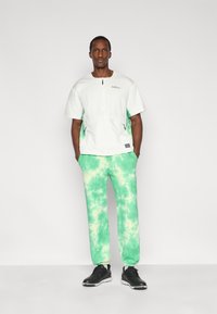 Λευκό πουκάμισο με φερμουάρ και πράσινες λεπτομέρειες και λογότυπο, συνδυασμένο με πράσινο παντελόνι tie-dye. Μαύρα αθλητικά παπούτσια ολοκληρώνουν την ενδυμασία.