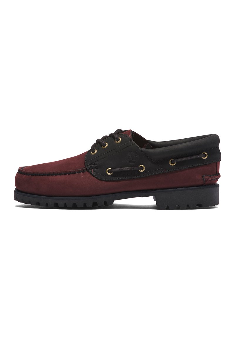 Zapato tipo barco de ante burdeos con detalles en cuero negro y ojetes de latón. Presenta una suela de goma texturizada y un detalle de costura a lo largo del borde.