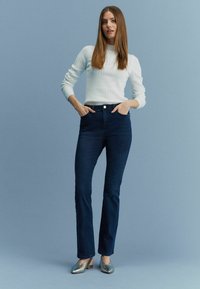 Jeans negri cu o croială dreaptă, asortați cu un pulover deschis cu fir răsucit și pantofi argintii cu toc mic, pe un fundal albastru.