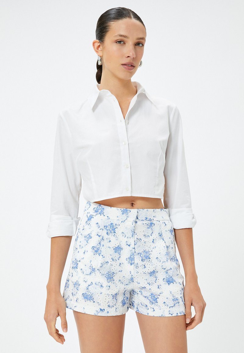 Witte cropped button-up shirt met lange mouwen, gecombineerd met high-waisted shorts met een blauw bloemenpatroon op een witte achtergrond.