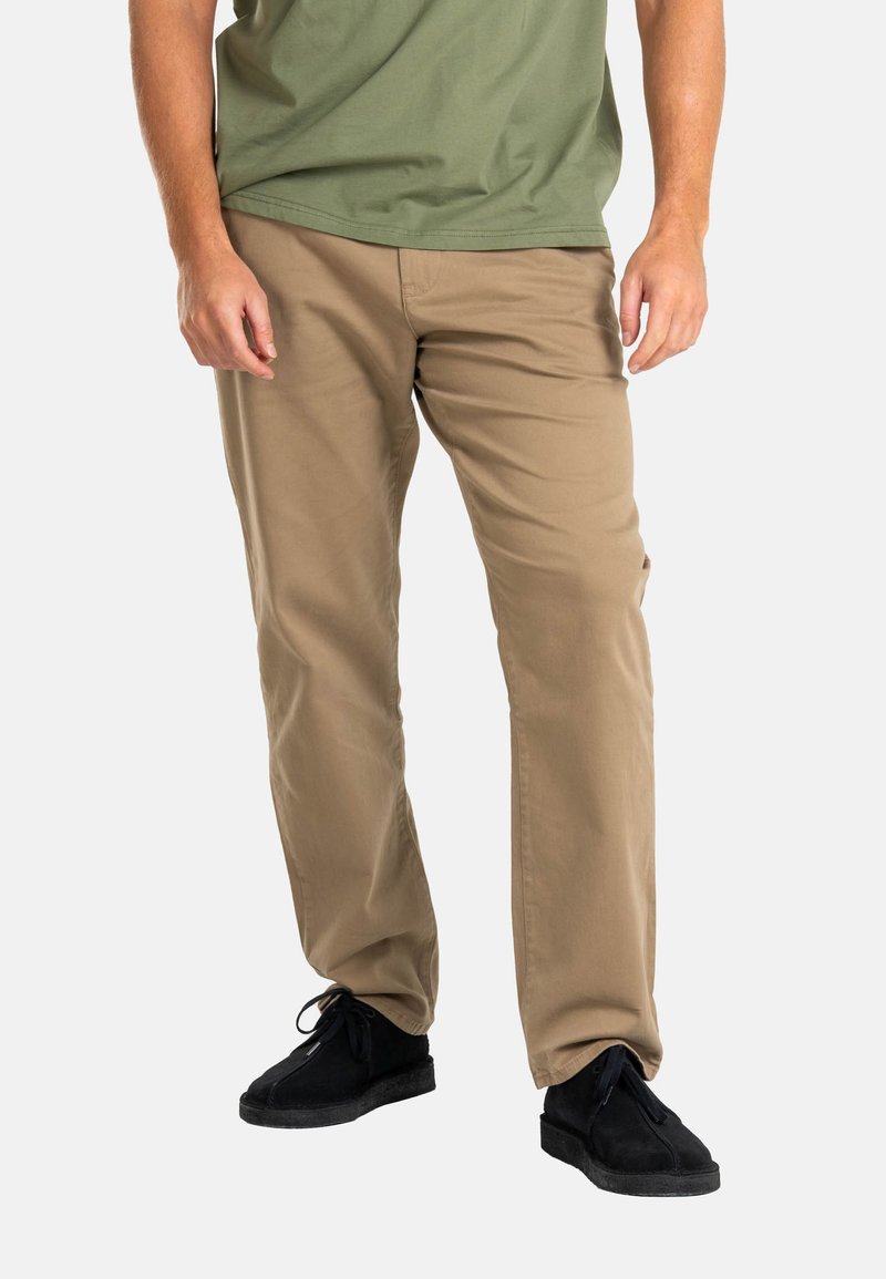 Beige cotton chinos with a straight fit, featuring side and back pockets, worn with black suede shoes. Simple, clean design.