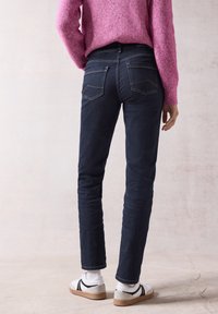 Jeans azul oscuro de corte slim, con dos bolsillos traseros que tienen un diseño de costura sutil. Combinados con zapatillas blancas y un suéter de punto rosa.