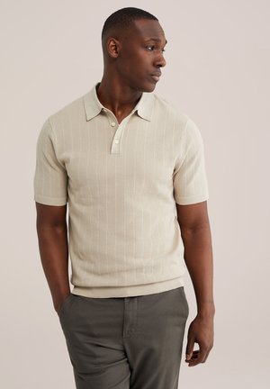 Mees seisab, parem käsi taskus, seljas lühikeste varrukatega beež kootud polo-särk ja tumedad hallid püksid, vaadates paremale.