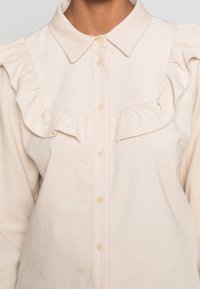 Helle beige Bluse mit Knopfleiste, gerüschter Front, kurzen Ärmeln und einem klassischen Kragen. Weiches Material mit subtiler Textur.