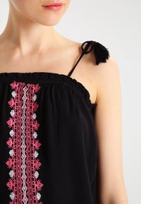 Top senza maniche nero con scollatura arricciata, caratterizzato da intricate ricami rosa e bianchi sulla parte anteriore e una nappina su una spallina.