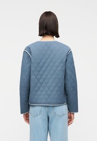 Άτομο με κοντά σκούρα μαλλιά που φοράει μπλε μπουφάν με καπιτονέ λεπτομέρειες και τζιν ψηλόμεσο με ανοιχτό μπλε χρώμα, κοιτάζοντας προς τα πίσω.