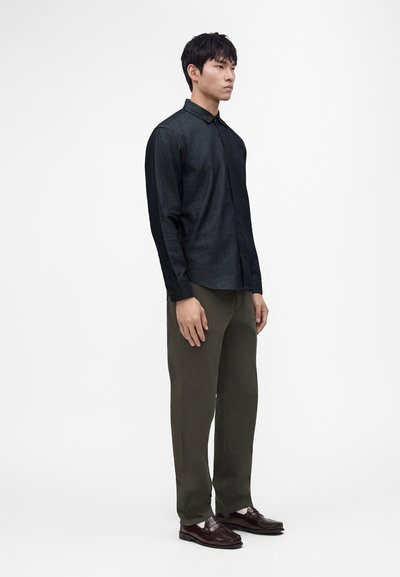 Homme portant une chemise sombre à boutons, un pantalon vert olive et des mocassins marron foncé, debout devant un fond blanc uni.