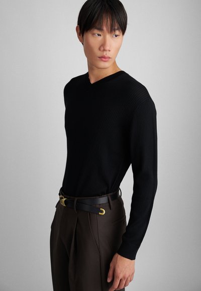 Schwarzer gerippter Pullover mit langen Ärmeln und V-Ausschnitt, kombiniert mit dunkelbraunen Hosen und einem schwarzen Gürtel mit goldener Schnalle.