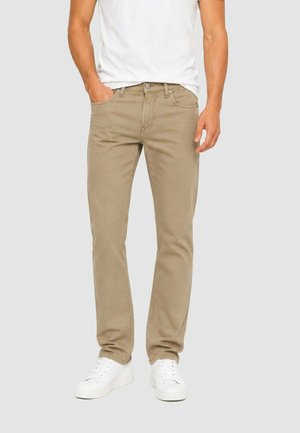 Uomo con pantaloni beige a gamba dritta, maglietta bianca e sneakers bianche, in piedi con una mano in tasca e l'altra di lato.
