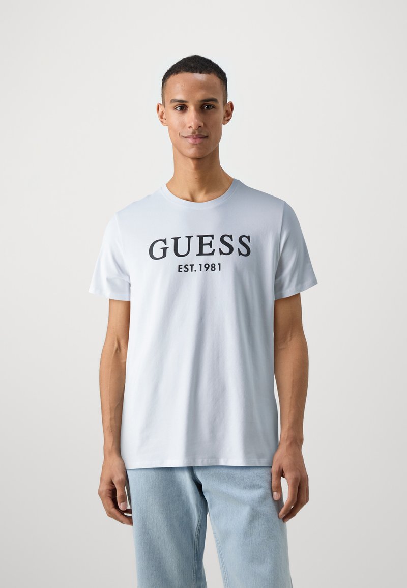 Světle modré bavlněné tričko s kulatým výstřihem, na přední straně s černým nápisem "GUESS EST.1981". Krátké rukávy a volný střih.