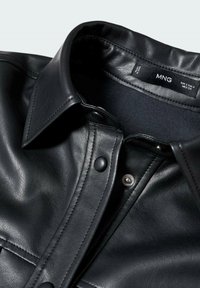 Mustapahkinapaita, jossa on muodollinen kaulus, nepparipainikkeet ja sileä rakenne; kauluksessa on "MNG"-merkki.