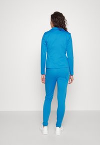 Modrý sportovní teplákový souprava z hladké, elastické látky; přiléhavá bunda s límcem a odpovídající slim kalhoty; bílé obuv.