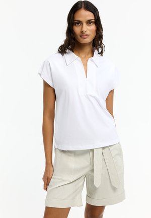 Frau trägt eine weiße kurzärmelige Bluse mit Kragen, kombiniert mit hellbeigen, hoch taillierten Shorts mit einem vorne gebundenen Stoffgürtel.