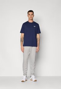 Marineblå t-shirt med korte ærmer og et lille logo, parret med lysegrå træningsbukser og hvide sneakers med sorte detaljer.